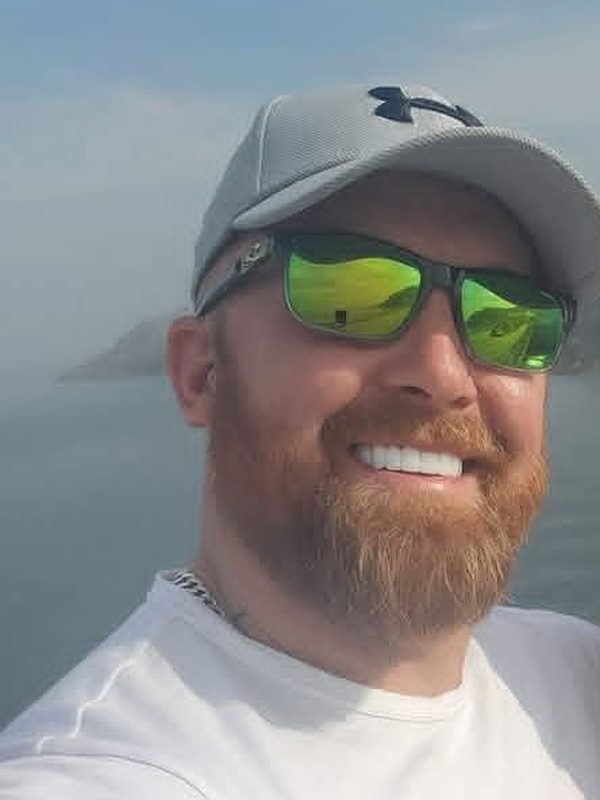 Greg Probert in a white T-shirt, grey cap and sunglasses, smiling on a boat with a headland behind him.