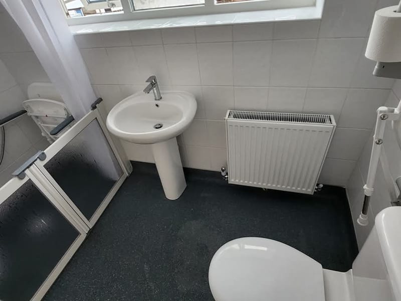 The rest of the same room: a pedestal basin with a lever tap, a panel radiator, slip-resistant safety flooring, and the half-height shower doors at the left.
