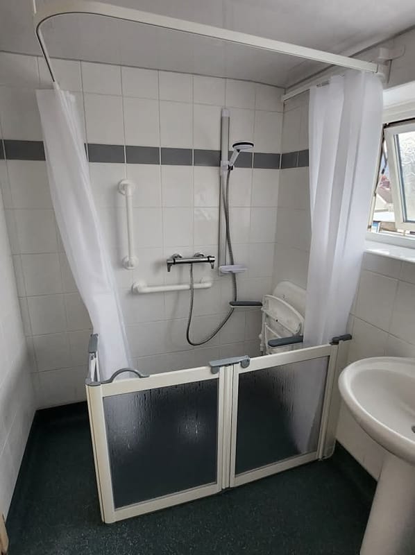 A level-access shower with half-height doors, a shower curtain on a rail, a thermostatic bar mixer, a wall-mounted fold-down seat, and a straight and an angled grab rail.