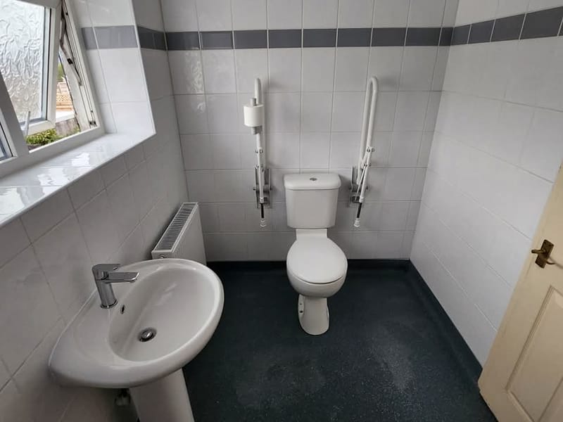 A close-coupled toilet with a drop-down grab rail on each side of it, beside a pedestal basin and a panel radiator.