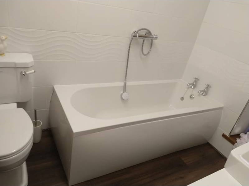 A white panelled bath with a thermostatic shower over it, next to a close-coupled toilet, on a dark wood-effect floor.