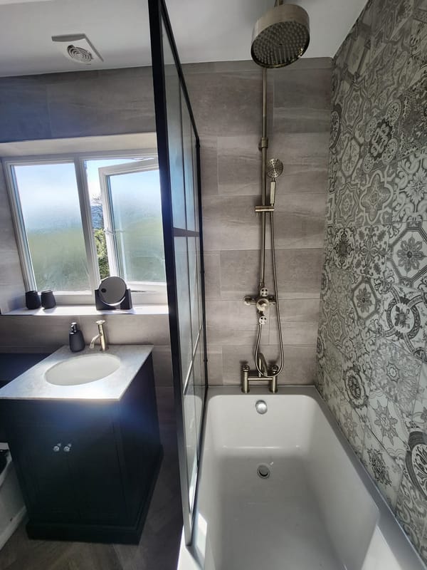 A bath with a brushed-brass riser-rail shower and a black-framed screen, against stone-effect tiling with a panel of patterned feature tiles, beside a dark vanity unit with a marble top.