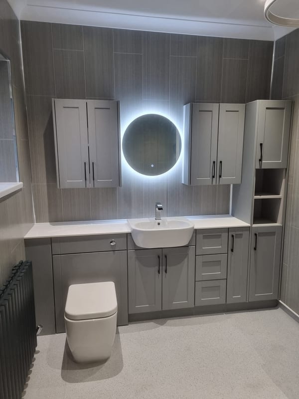A run of grey fitted bathroom furniture: a basin set into a white worktop, a back-to-wall toilet, wall cabinets either side of a round illuminated mirror, and an anthracite column radiator at the end.