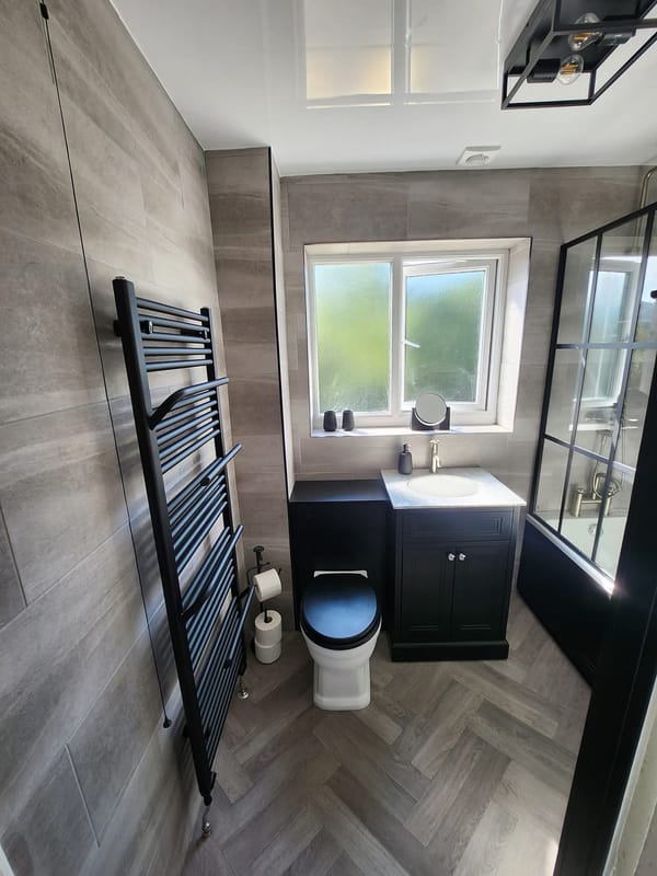 The same room from the door: a black towel radiator, a dark vanity unit and toilet unit under the window, and a herringbone wood-effect floor.