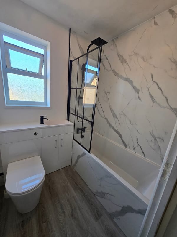 A bathroom in marble-effect panelling with black fittings: a bath with a black-framed screen and a square rainfall shower head, and a white unit carrying the basin and a back-to-wall toilet.