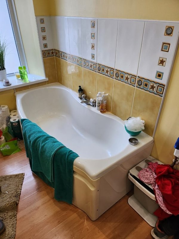 The old bathroom: a white bath against yellow and white tiles with a patterned border, on a wood-effect floor.