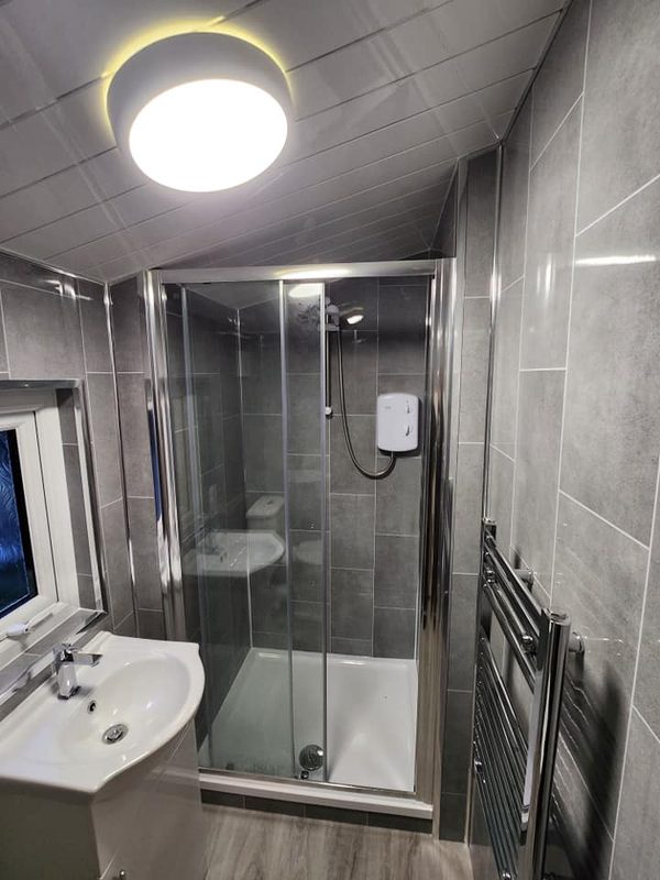 The same room from the same spot, finished: grey panelled walls, a white panelled ceiling with a round light, a shower cubicle, a basin on a white unit and a chrome towel radiator.