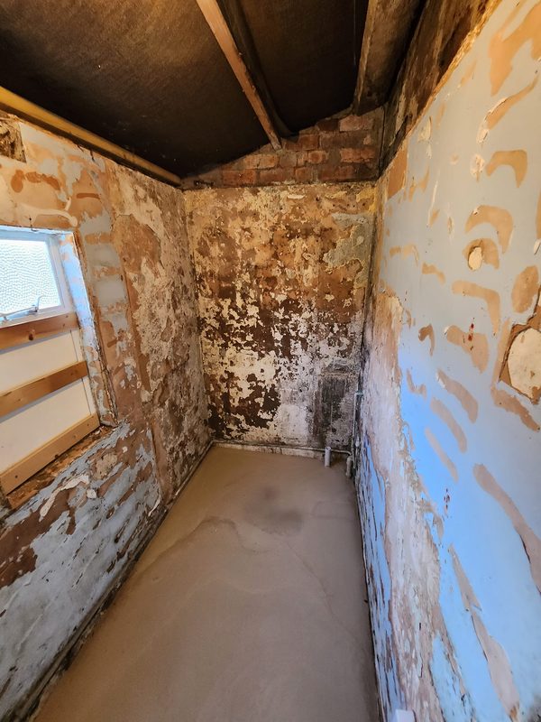 The coal shed stripped back to bare brick and old plaster, with a bare bulb, a boarded roof and a small window on the left.