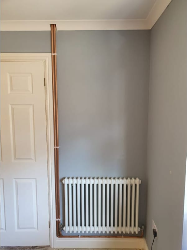 A white column radiator fitted to a grey wall, fed by neatly clipped copper pipework running up beside the door frame.