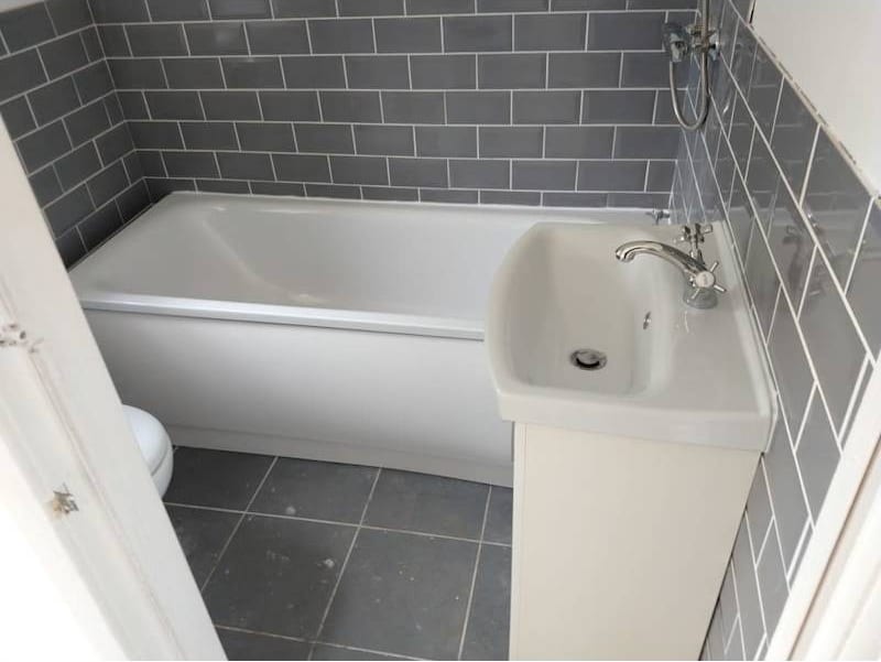 A compact bathroom with grey metro tiles, a bath and a basin mounted on a unit over the toilet.