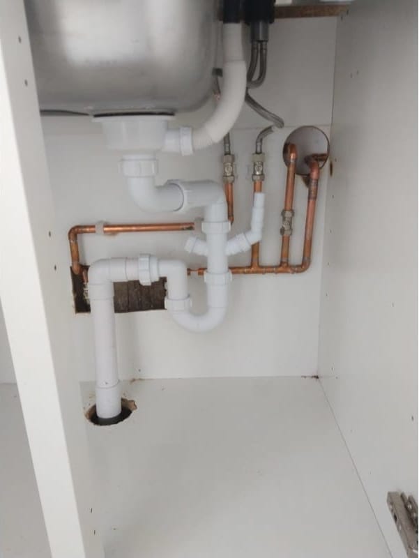 Inside a kitchen sink unit: white waste pipes and a trap under a stainless steel sink, with copper hot and cold feeds on isolation valves.