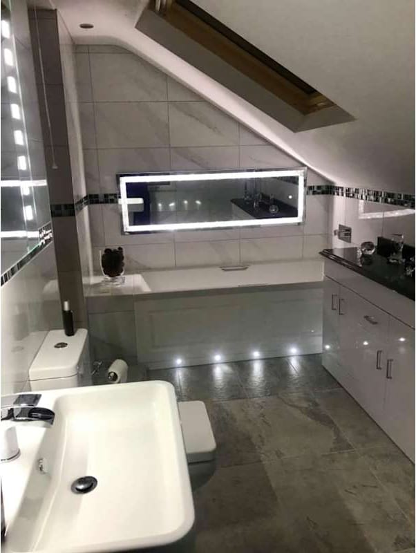 A loft bathroom with marble-effect tiling, a bath under the eaves, an illuminated mirror and gloss white units.
