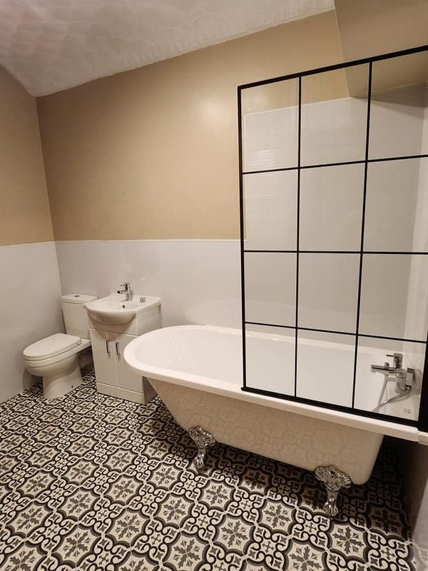 A white roll-top bath on feet with a black-framed shower screen, beside a toilet and basin, on a black and white patterned floor.