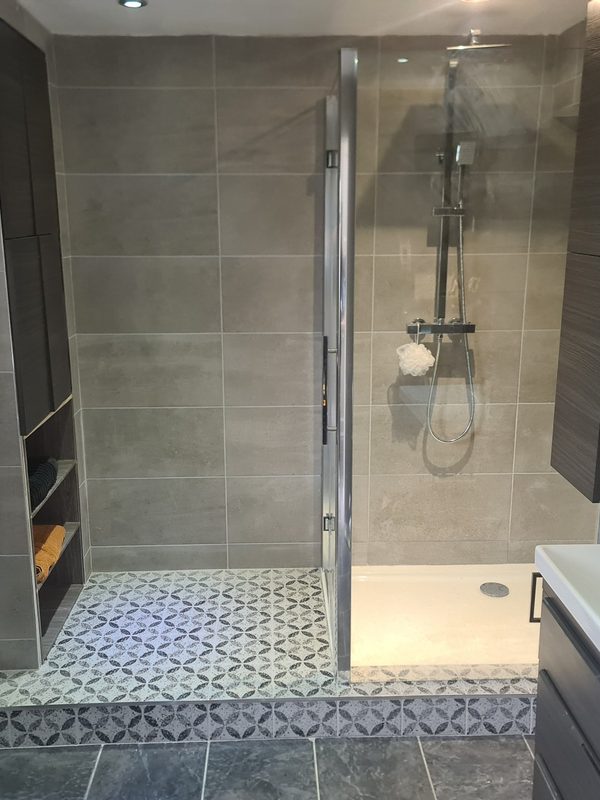 A shower enclosure with a glass door and a thermostatic riser-rail shower against large grey tiles, above a floor of blue and white patterned tiles.