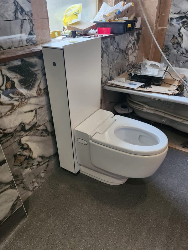 A wall-hung shower toilet fitted against marble-effect panelling, its cistern concealed in the slim white unit beside it, in a bathroom still being worked on.