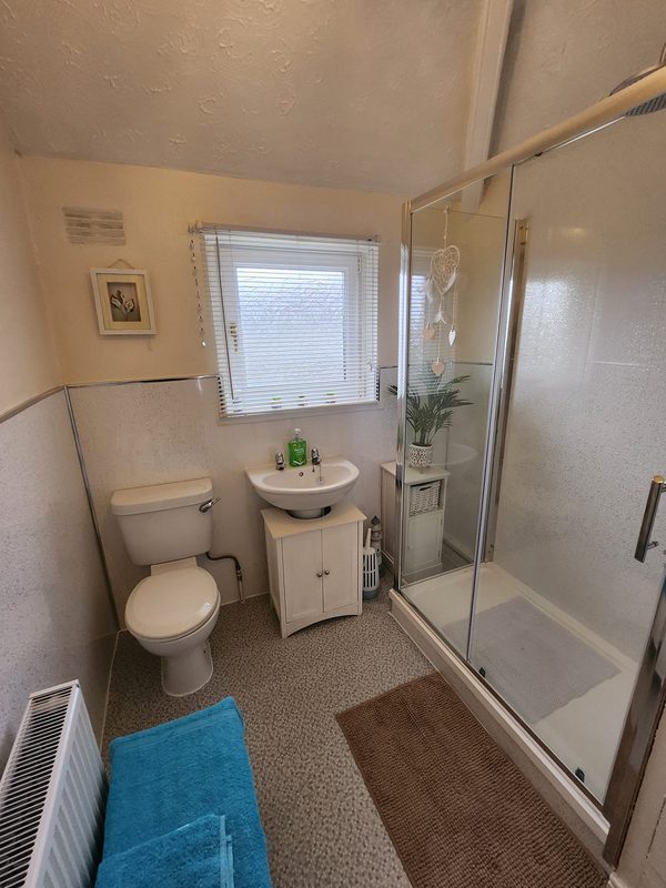 The same corner: a tall glass shower cubicle where the bath was, sparkly white wall panels, the basin unit and toilet under the window.