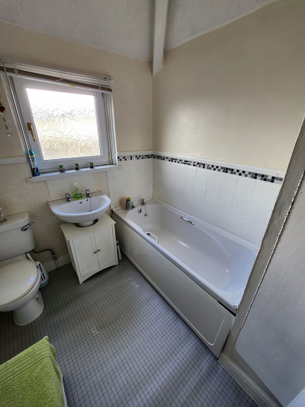The old bathroom under a small window: a white bath with a mosaic border, a basin on a small unit and a toilet, on a grey floor.