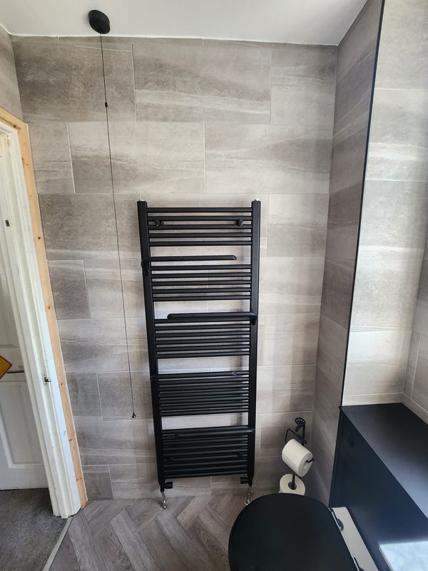 A tall matt black ladder towel radiator on grey stone-effect wall panels.