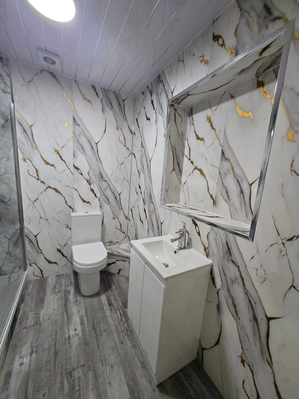 The same room from the doorway: marble-effect panels on every wall and a panelled ceiling, a toilet and a white vanity basin under a recessed mirror, and a grey wood-effect floor.